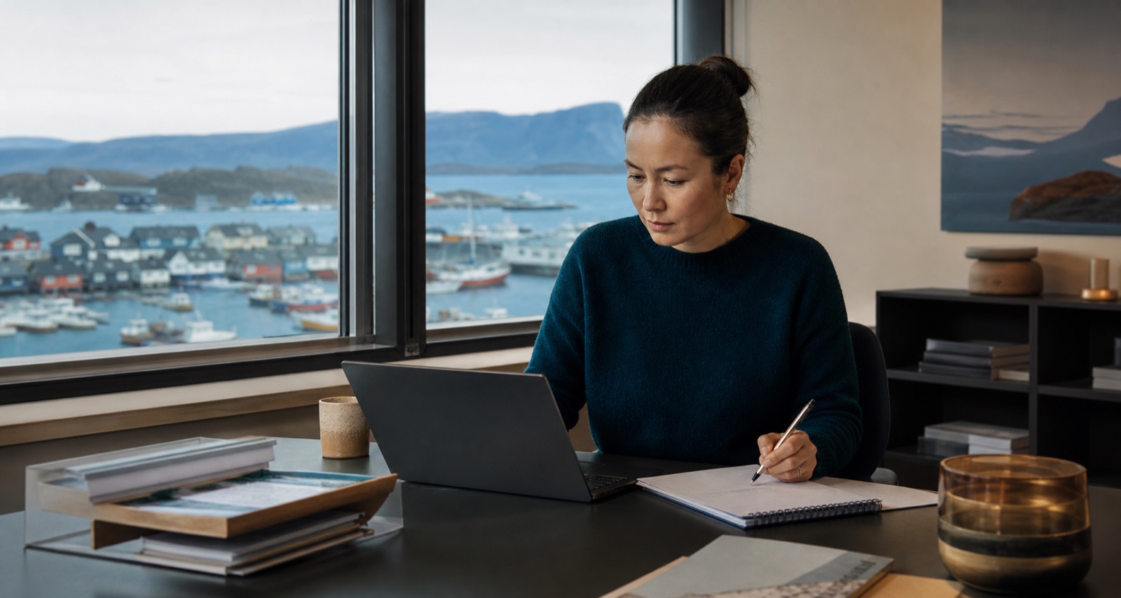 Lønadministrator arbejder ved en computer på et kontor med udsigt over en grønlandsk havn.